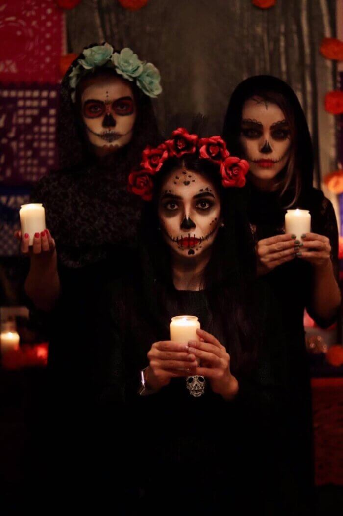 Tři ženy v kostýmu La Catrina s malovanými lebkami a svíčkami během oslav Día de los Muertos v Mexiku.