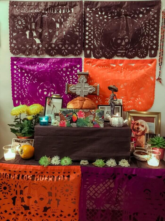 Černý, fialový a oranžový papel picado s motivem Día de los Muertos a malý oltář s ofrenda ke dni mrtvých s křížem, pan de uerto a keramickou sochou catriny, květinami a fotkami osob a pejska. 