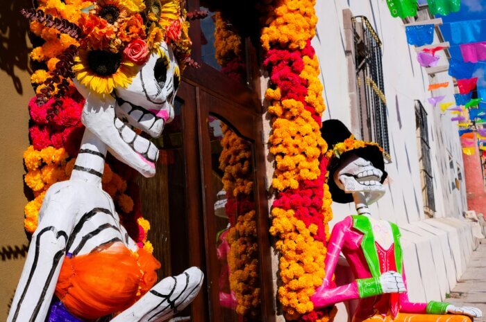 Ulice v Mexiku vyzdobená na Día de los Muertos, s postavou la Catrina a el Catrín, s květy cempasúchil a papel picado.