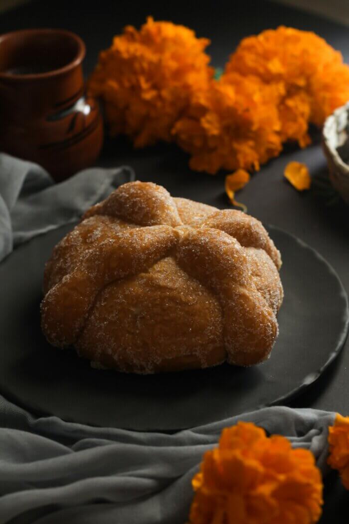 Pan de muerto na černém talíři na stole který je ozdoben květinami cempasúchil.