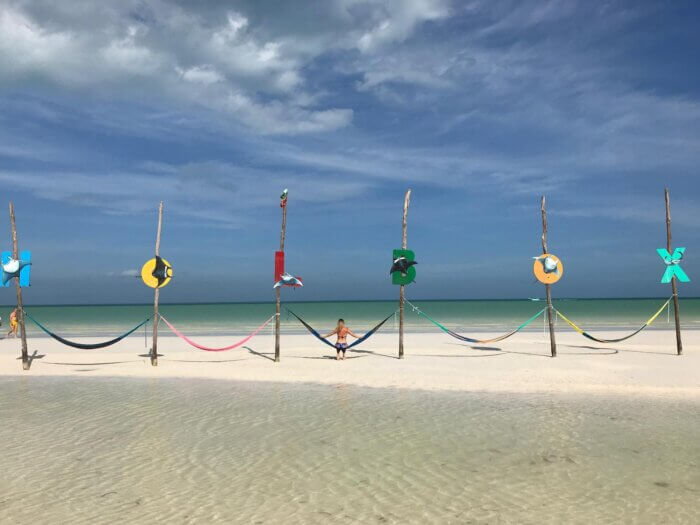 Osoba sedící na pláži mezi barevnými hamakami pověšenými na dřevěných panelech s výhledem na tyrkysové moře a modrou oblohu, každý sloup má na sobě barevné písmeno a dohromady tvoří nápis HOLBOX.