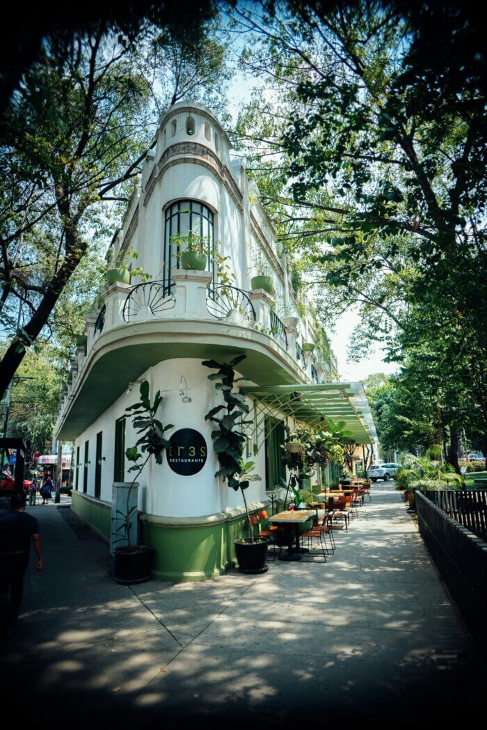Rohový dům v art deco stylu se zelenými akcenty a restaurací TRES v čtvrti Condesa, Mexico City, obklopený stromy a stolky na chodníku.