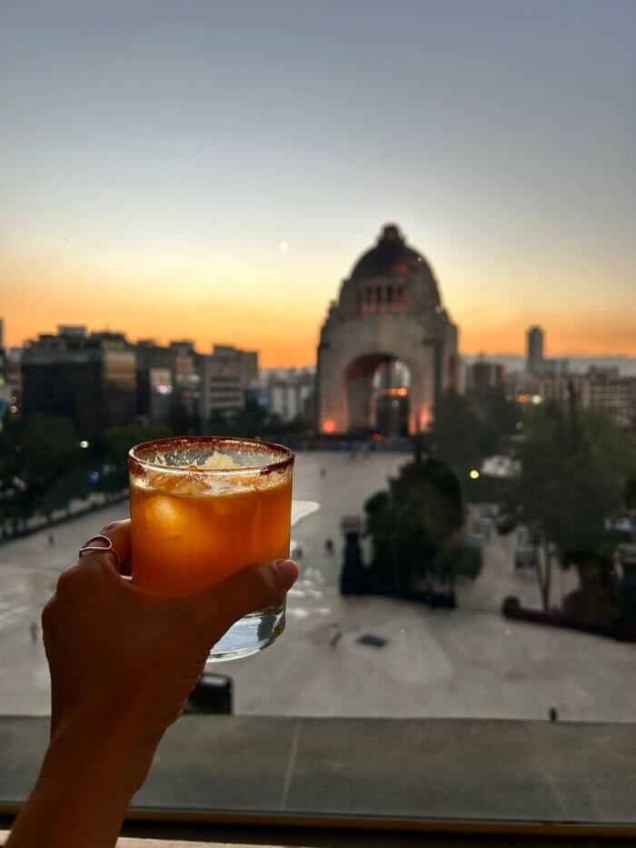 Koktejl s mezcalem na střešní terase Terraza Cha Cha Chá s výhledem na Monumento a la Revolución v Mexico City při západu slunce.