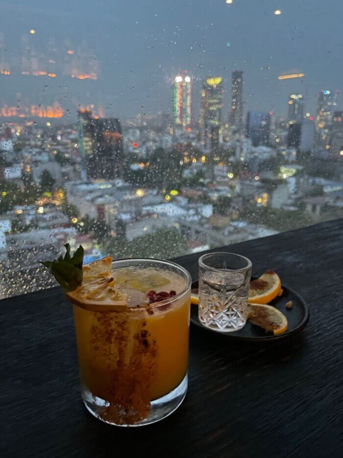 Koktejl a panák mezcalu na stole rooftop baru Supra Roma s výhledem na rozsvícené mrakodrapy Mexico City po setmění.