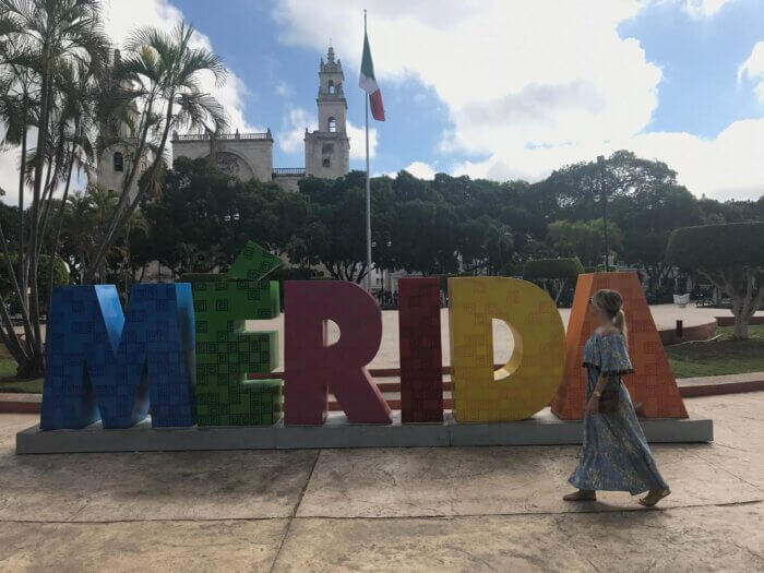 Žena v modrých šatech prochází kolem barevného nápisu 'MÉRIDA' na náměstí v Méridě, Mexiko, s historickou budovou v pozadí.